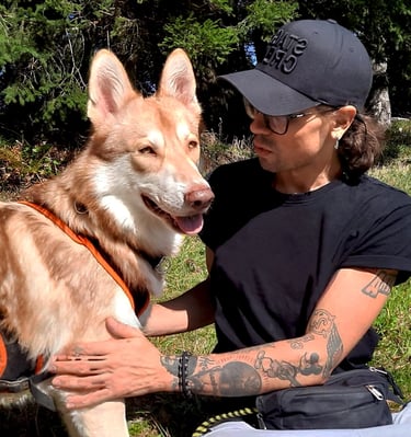 Mann in einem schwarzen Hemd und einer schwarzen Baseballkappe sitzt mit einem braunen Hund im Gras