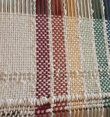 a loom with an unfinished hand-woven towel in muted rainbow pride flag colours