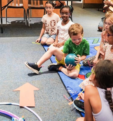 a group of children sitting on a rug in a classroom passing a weighted ball STEM