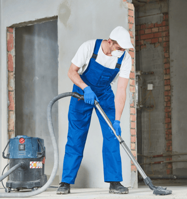 Trabajador con mono de tirantes azul, camiseta blanca y gorra, limpiando en una obra con aspirador.