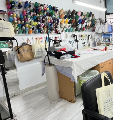 Vista general de un taller de costura con varias máquinas de coser, mesa de corte, rollos de tela, h