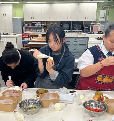 同學們喺設備齊全嘅廚房中專注裝飾甜點，使用唧花袋為 泡芙加上糖霜和士多啤梨裝飾，營造出互動學習嘅熱鬧氛圍