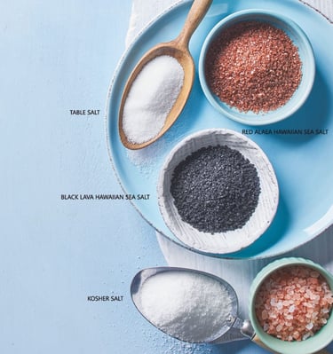 Various salts in bowls and spoons on a blue surface: table, kosher, red and black Hawaiian sea salt.