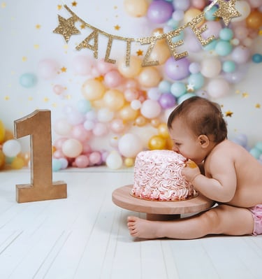 Baby girl celebrating a first birthday cake smash with pink frosting and pastel balloon backdrop.