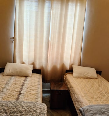 Two single beds with beige patterned bedding and white pillows in a cozy bedroom with light brown curtains.