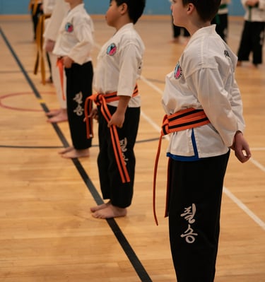 Young children in martial arts uniforms with orange belts standing in a row during a taekwondo class.