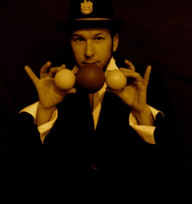 a man in a suit and hat with two hands up to the camera holding juggling balls