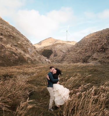 couple in a field of grass with wind turbine in the background at makara beach