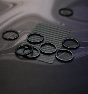 Small black circular split rings and metal fasteners for EDC gear on a carbon fiber mat.