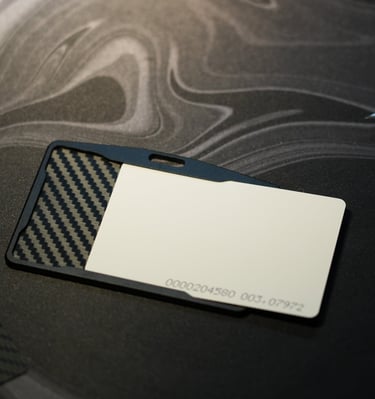 A carbon fiber ID badge holder with a blank white plastic card on a dark marble desk.