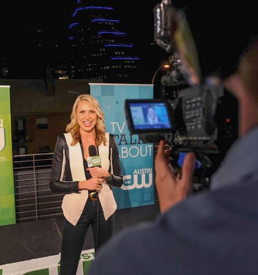 CW Star smiles on-camera, recording segment with pro gear against branded backdrop and city lights.