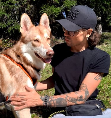 photo of a man wearing a black baseball cap and glasses sitting on the grass with a large brown dog
