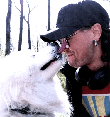 a photo of a man wearing a black baseball cap and glasses being licked in the face by white dog