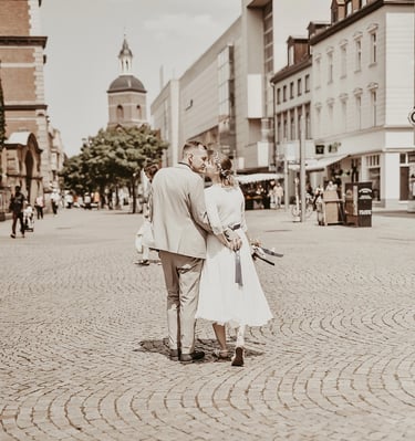 Ein Hochzeitsfoto, das ein Paar von hinten beim Küssen zeigt, während sie durch eine gepflasterte Straße gehen. Der Mann träg