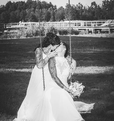  Dieses Schwarz-Weiß-Bild zeigt zwei Bräute in weißen Hochzeitskleidern, die sich küssen, während sie auf einer Schaukel sitz
