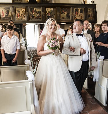  Ein Bild, das eine lächelnde Braut zeigt, die von einem Mann, wahrscheinlich ihrem Vater, den Gang einer Kirche entlanggefüh
