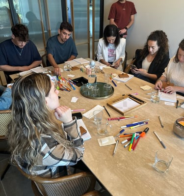 קבוצת משתתפים בסדנה יושבים סביב שולחן, עובדים על ארנקי כוחות העל שלהם עם כלי כתיבה ויצירה.