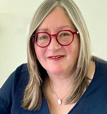 Professional portrait of a smiling woman with blonde hair wearing stylish red glasses and a blue v-neck top.