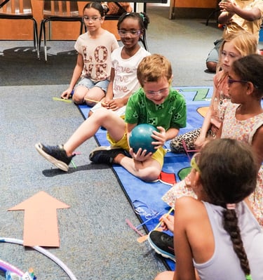 a group of children sitting on a rug in a classroom passing a weighted ball STEM
