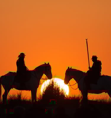 Silhouette of two Camargue horse riders at sunset with a vibrant orange sky background.