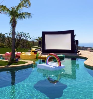 A swimming pool is surrounded by inflatable toys under a clear sky, with a large outdoor screen rental and speakers set 
