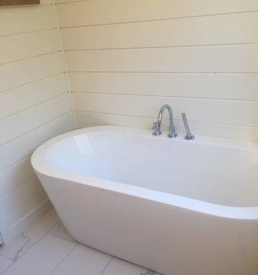 Modern white freestanding soaking bathtub with chrome faucet against cream shiplap walls.