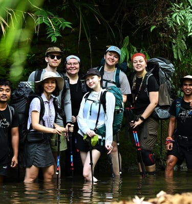 A diverse group of hikers and guides wading through a jungle river during a tropical trekking expedition.