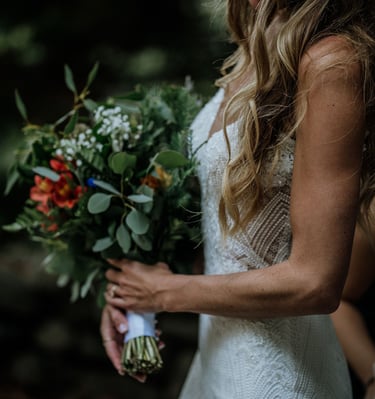 A smiling bride in a lace wedding dress holding a rustic wildflower bouquet with greenery.