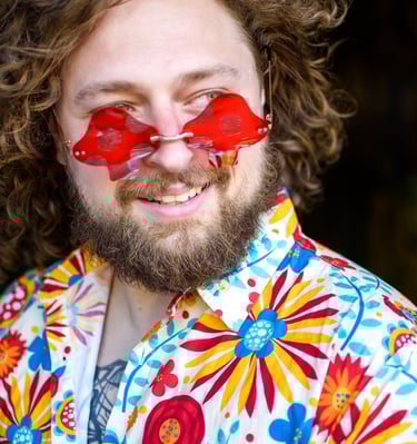 A smiling man with a beard wearing mushroom-shaped red sunglasses and a colorful floral shirt