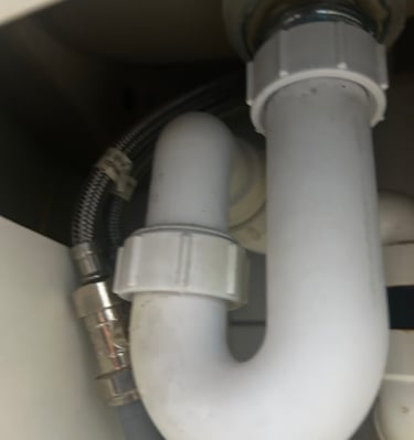 Under-sink plumbing showing a white plastic P-trap pipe and braided metal water supply line.