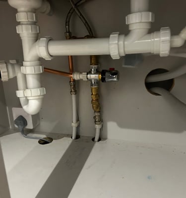 Under-sink plumbing with white PVC drain pipes, copper lines, and a thermostatic mixing valve.