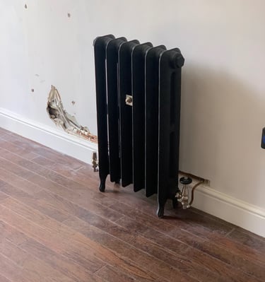 Black traditional cast iron radiator installed against a white wall with exposed copper piping.