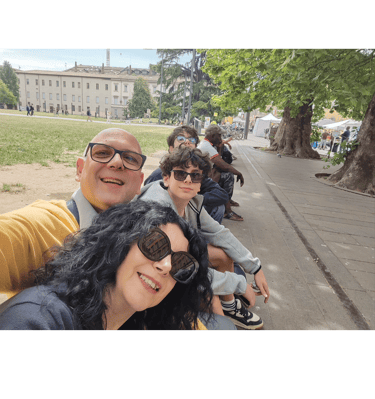 A smiling family takes a group selfie outdoors on a sunny day in an urban park square.