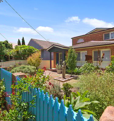 Front view of Marche Home Stay with a colourful garden, blue picket fence, and welcoming landscaped entrance.
