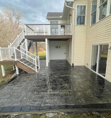 New dark grey stamped concrete patio installed under a two-story residential deck with white railings.