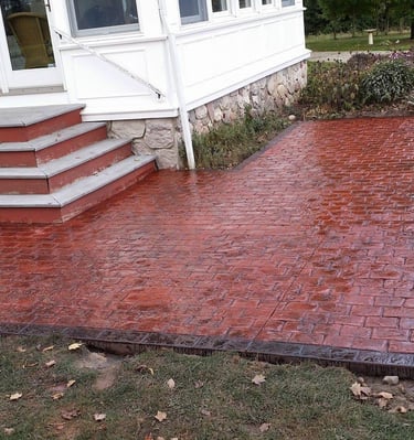Red stamped concrete patio with a decorative brick pattern installed next to a house's wooden stairs.