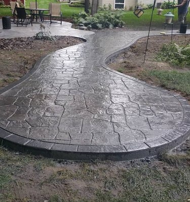 Curved dark gray stamped concrete walkway and patio in a residential backyard landscape.
