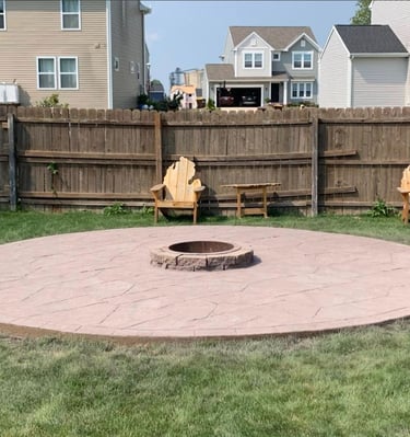 Circular stamped concrete patio with central fire pit and Adirondack chairs in a fenced backyard.