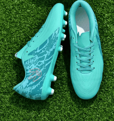 Un par de botas de fútbol de color azul turquesa sobre un campo de césped artificial verde.