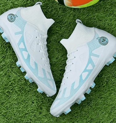 Un par de botas de fútbol de caña alta en color blanco y azul claro, sobre cesped verde.