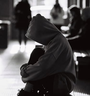 A person wrapped in an oversized hoodie, sitting alone in a public space