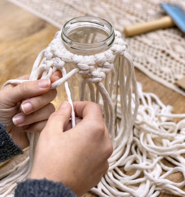  Manos atando nudos para una tapa de frasco de macramé hecha a mano usando cordón de algodón blanco 