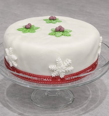 Festive white fondant Christmas cake on a glass stand decorated with holly and snowflakes.
