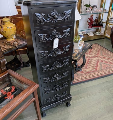 Elegant black lingerie chest with ornate floral wood carvings on seven drawers in an antique shop.