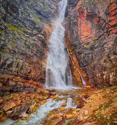 Scenic mountain waterfall cascading down colorful rocky cliffs into a clear stream.