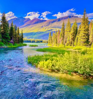Clear blue river flowing through a lush evergreen forest with majestic mountains under a bright sky.
