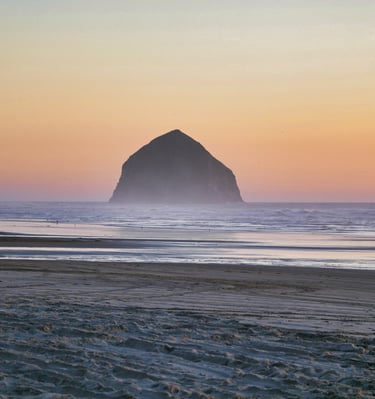 A wide sandy beach at sunset featuring the iconic Haystack Rock monolith off the Oregon coast.