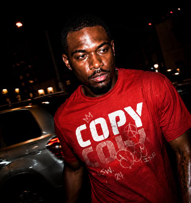 A dark, intense portrait of a man in a red "COPY" t-shirt, sweat glistening on his face, in a nighttime urban setting.