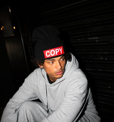 A man in a grey hoodie and black COPY logo beanie sits against a dark shutter door.