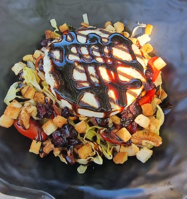 Warm goat cheese salad with balsamic glaze, crunchy croutons, and walnuts in a black bowl.